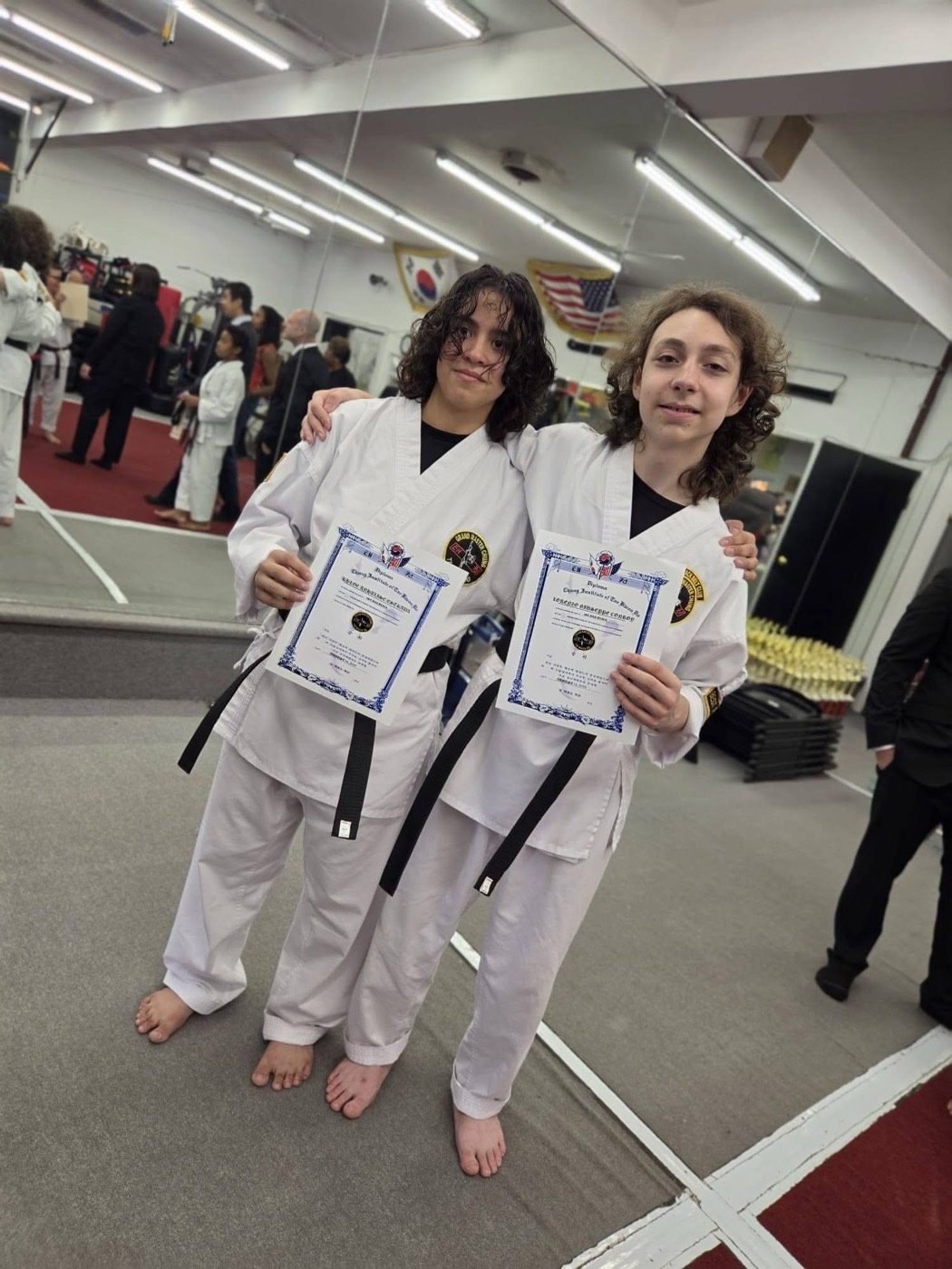 Two students in white uniforms with black belts, holding certificates at the school.